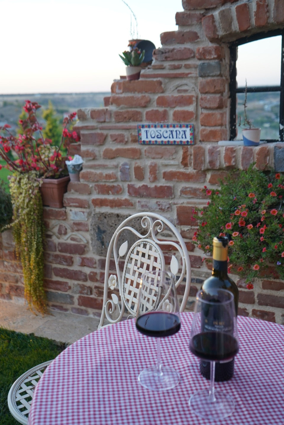 Două pahare de vin roșu pe o masă în carouri, lângă zidul de cărămidă cu inscripția Toscana